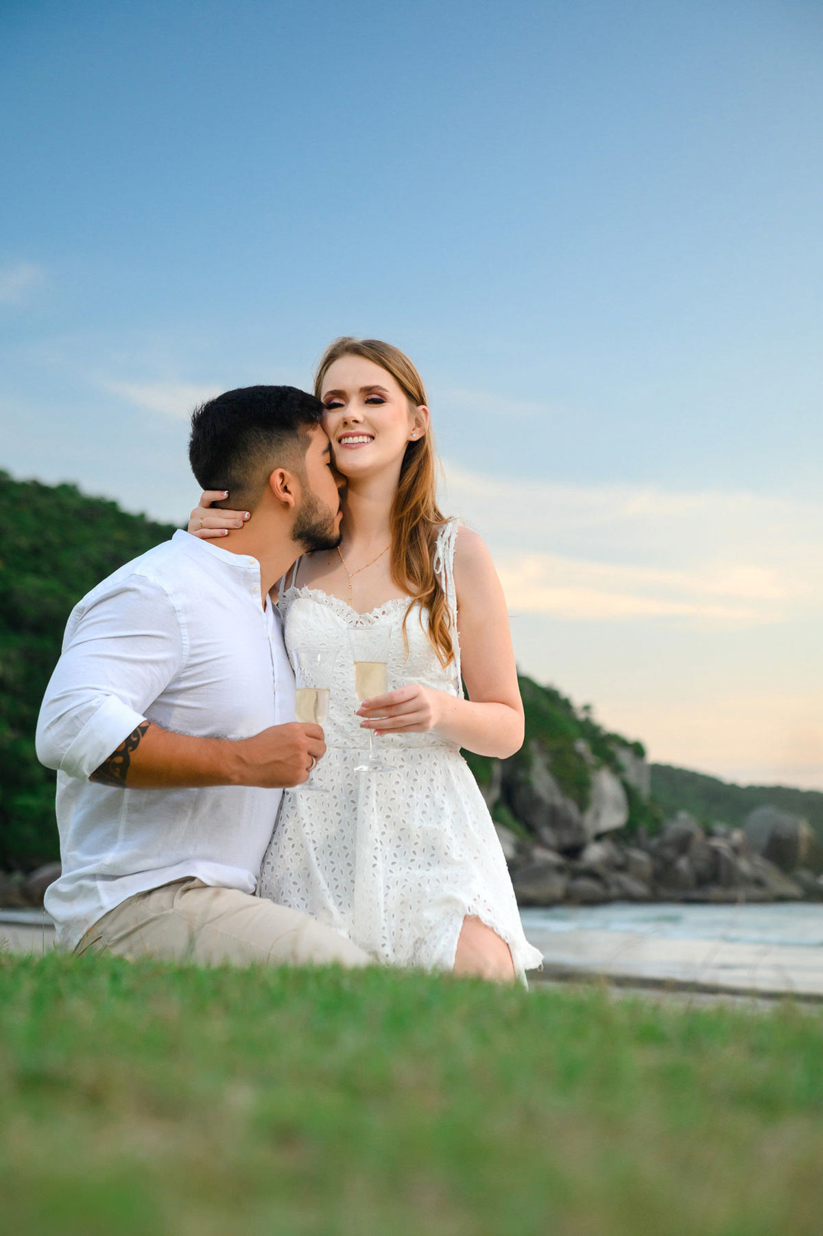 Casal brindando com taças na beira do mar ao amanhecer na praia de Bombas em Bombinhas, com nuvens dramáticas no horizonte.