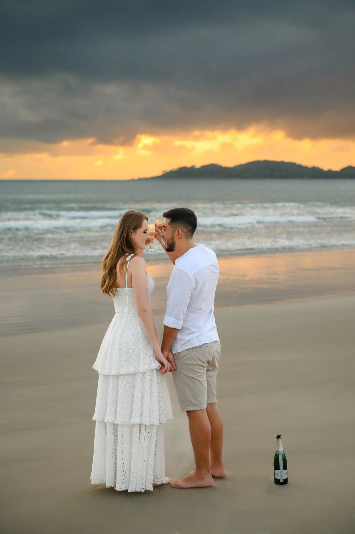 Casal brindando com taças na beira do mar ao amanhecer na praia de Bombas em Bombinhas, com nuvens dramáticas no horizonte.