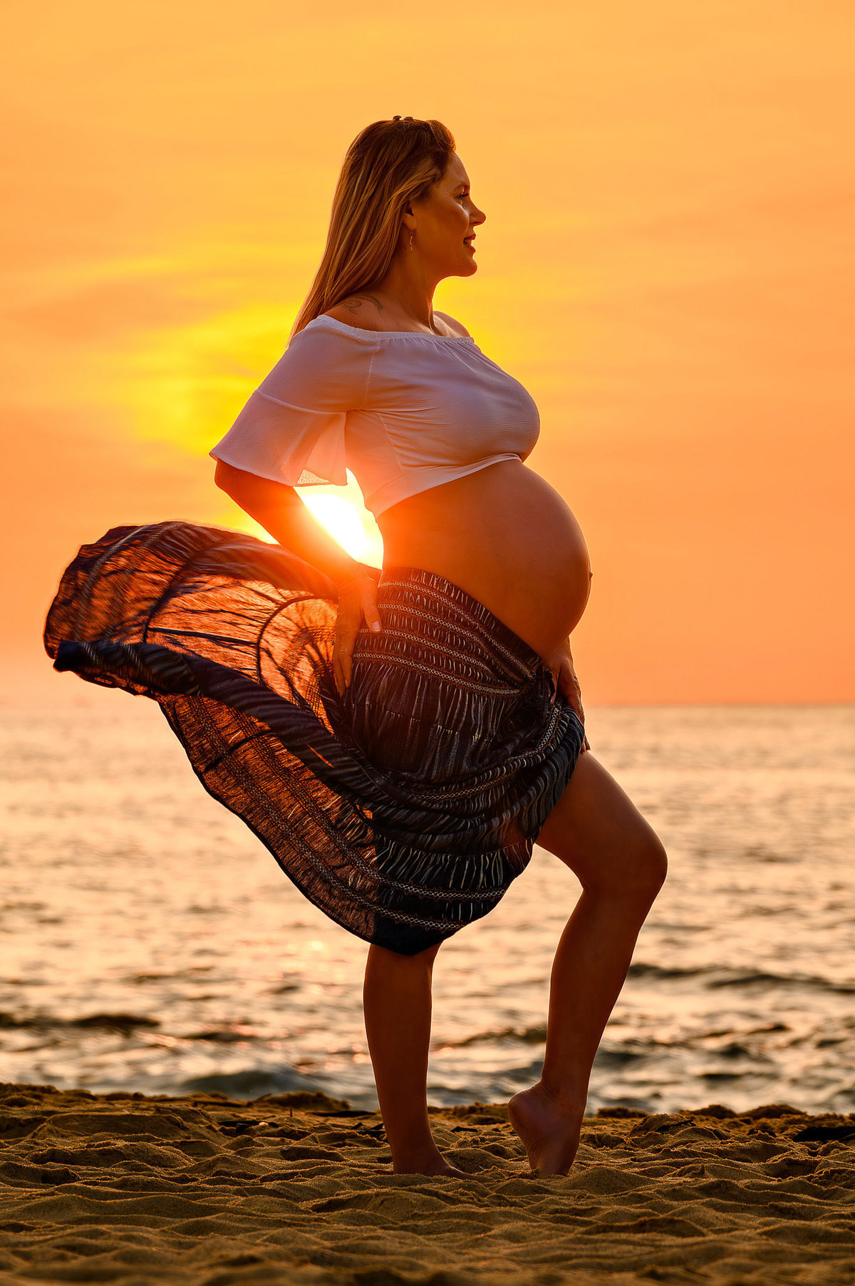 Silhueta de gestante ao amanhecer na praia do Estaleirinho, em Balneário Camboriú SC, com o sol nascendo ao fundo e o vestido esvoaçando