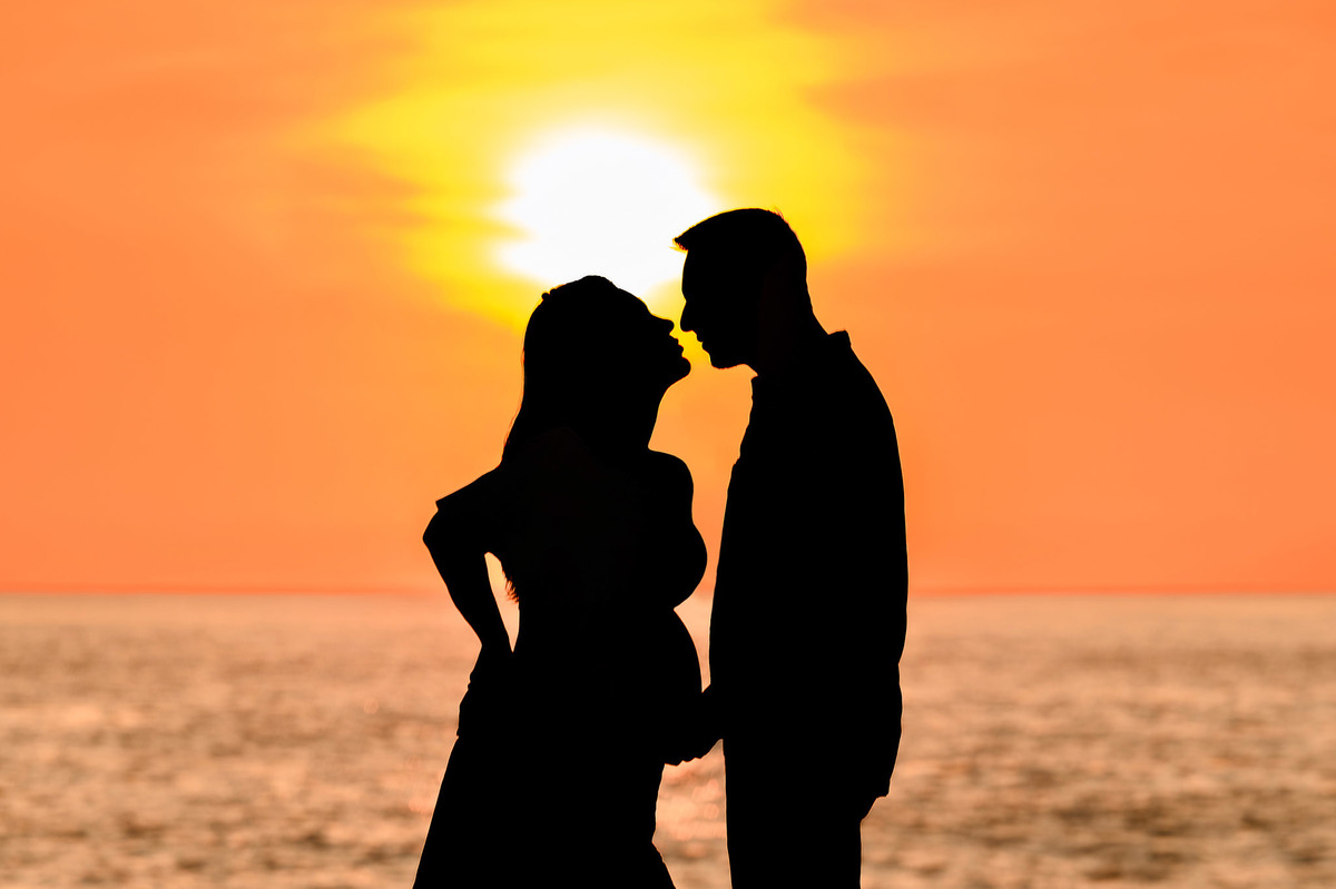 Silhueta de gestante e companheiro na praia ao amanhecer em Balneário Camboriú, criando uma cena romântica com o nascer do sol ao fundo.