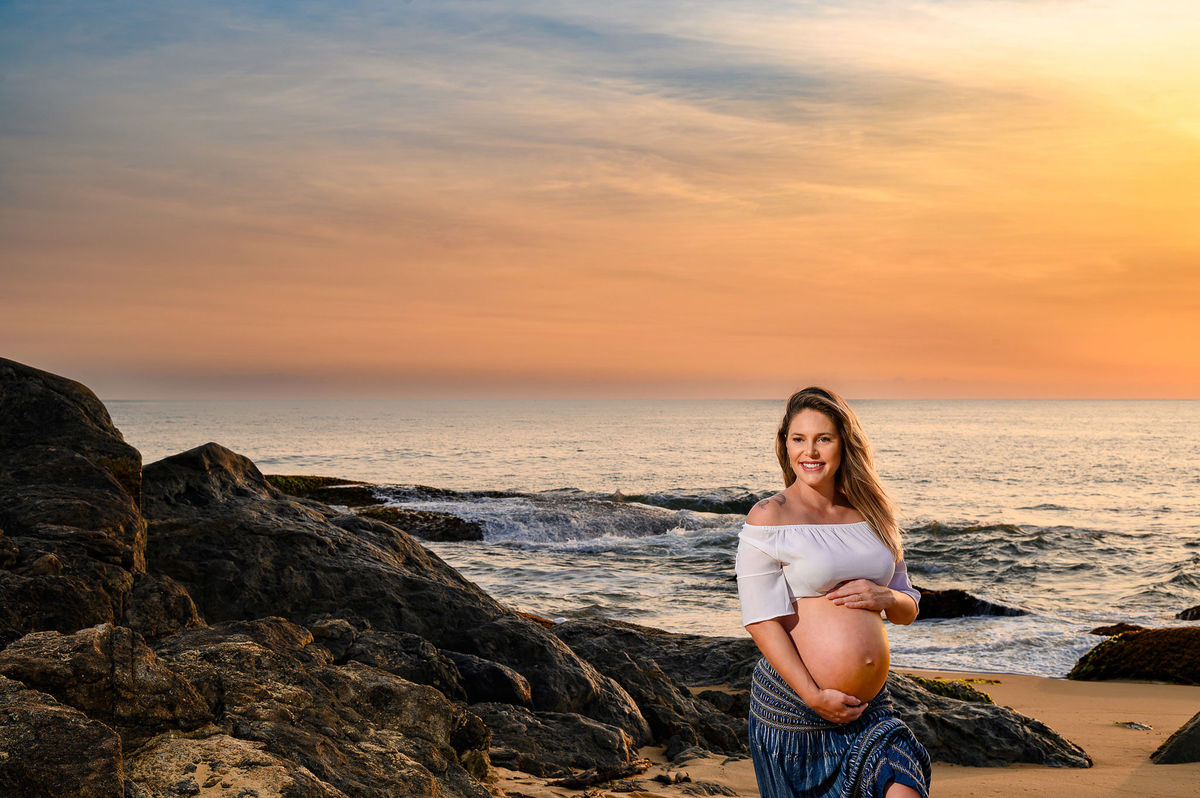 Fotografia de gestante ao amanhecer na praia do Estaleirinho, em Balneário Camboriú SC, com o mar ao fundo e céu em tons dourados