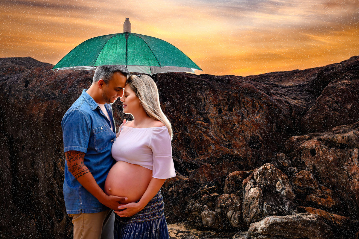 Casal em ensaio gestante ao amanhecer na praia em Balneário Camboriú segurando guarda-chuva, com luz suave e clima intimista.