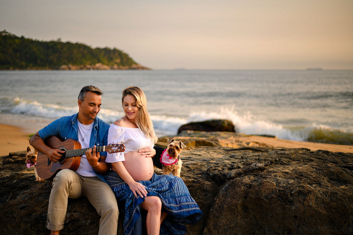 Ensaio gestante ao amanhecer na praia em Balneário Camboriú com casal sentado nas pedras, violão e cachorrinhas, luz suave do nascer do sol.