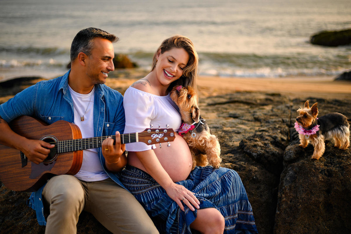Ensaio gestante ao amanhecer na praia em Balneário Camboriú com casal sentado nas pedras, violão e cachorrinhas, luz suave do nascer do sol.