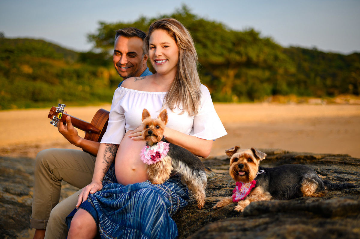 Ensaio gestante ao amanhecer Gestante sentada nas pedras da praia ao amanhecer em Balneário Camboriú ao lado do companheiro tocando violão, com cachorrinhas participando do ensaio.