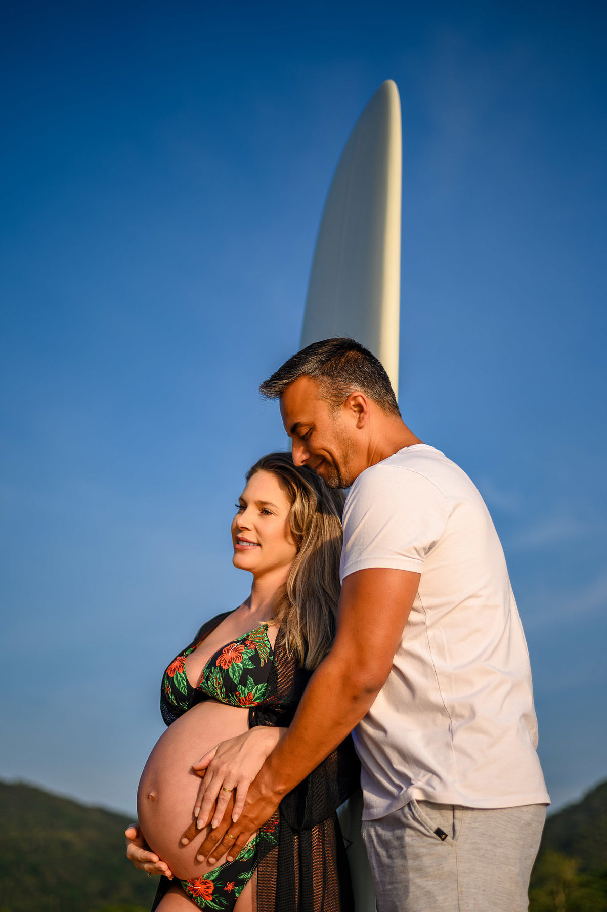 Casal em ensaio gestante na praia ao amanhecer em Balneário Camboriú com prancha de surf, em clima íntimo e natural.