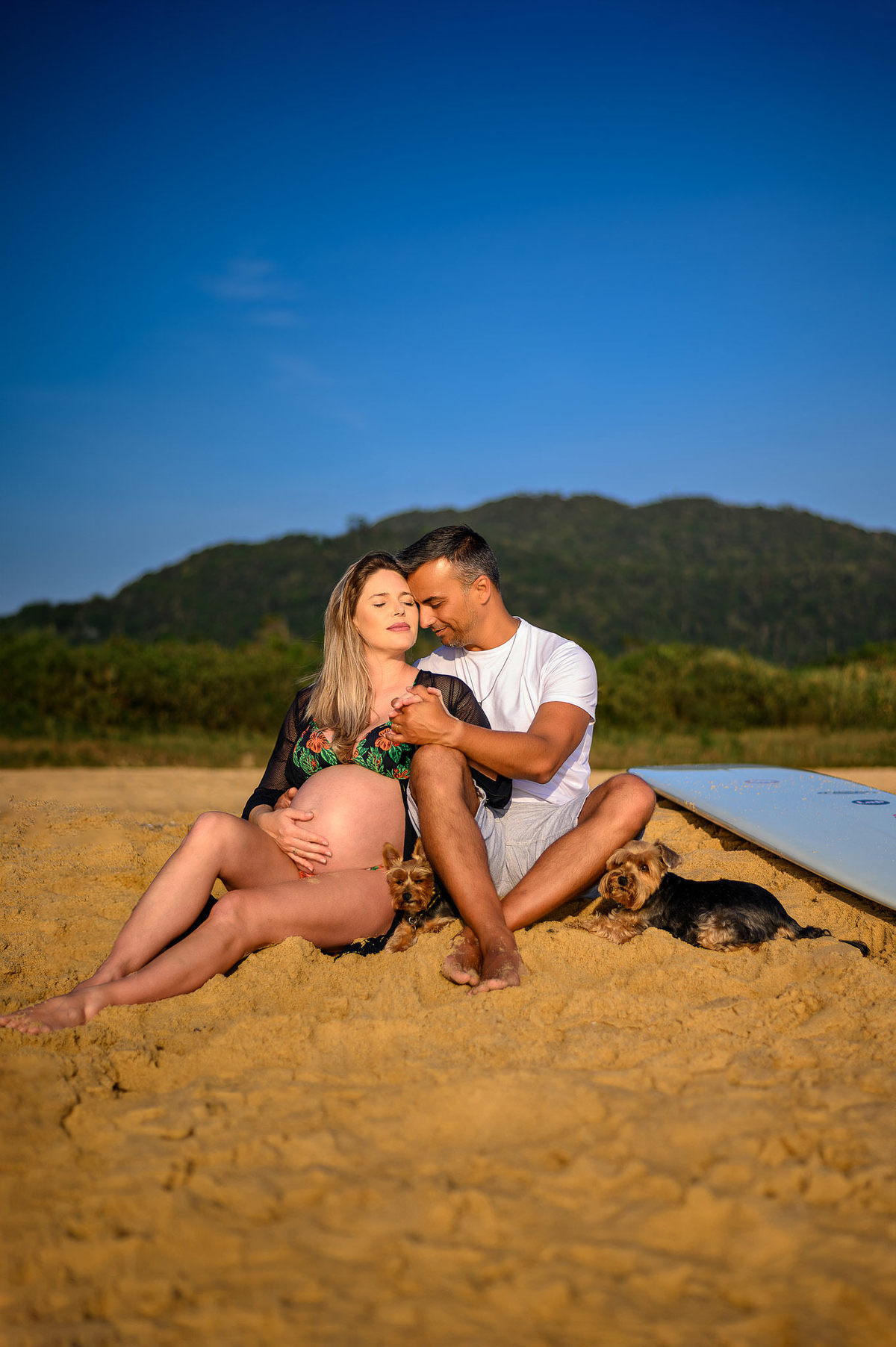 Casal em ensaio gestante na praia ao amanhecer em Balneário Camboriú com prancha de surf, em clima íntimo e natural.