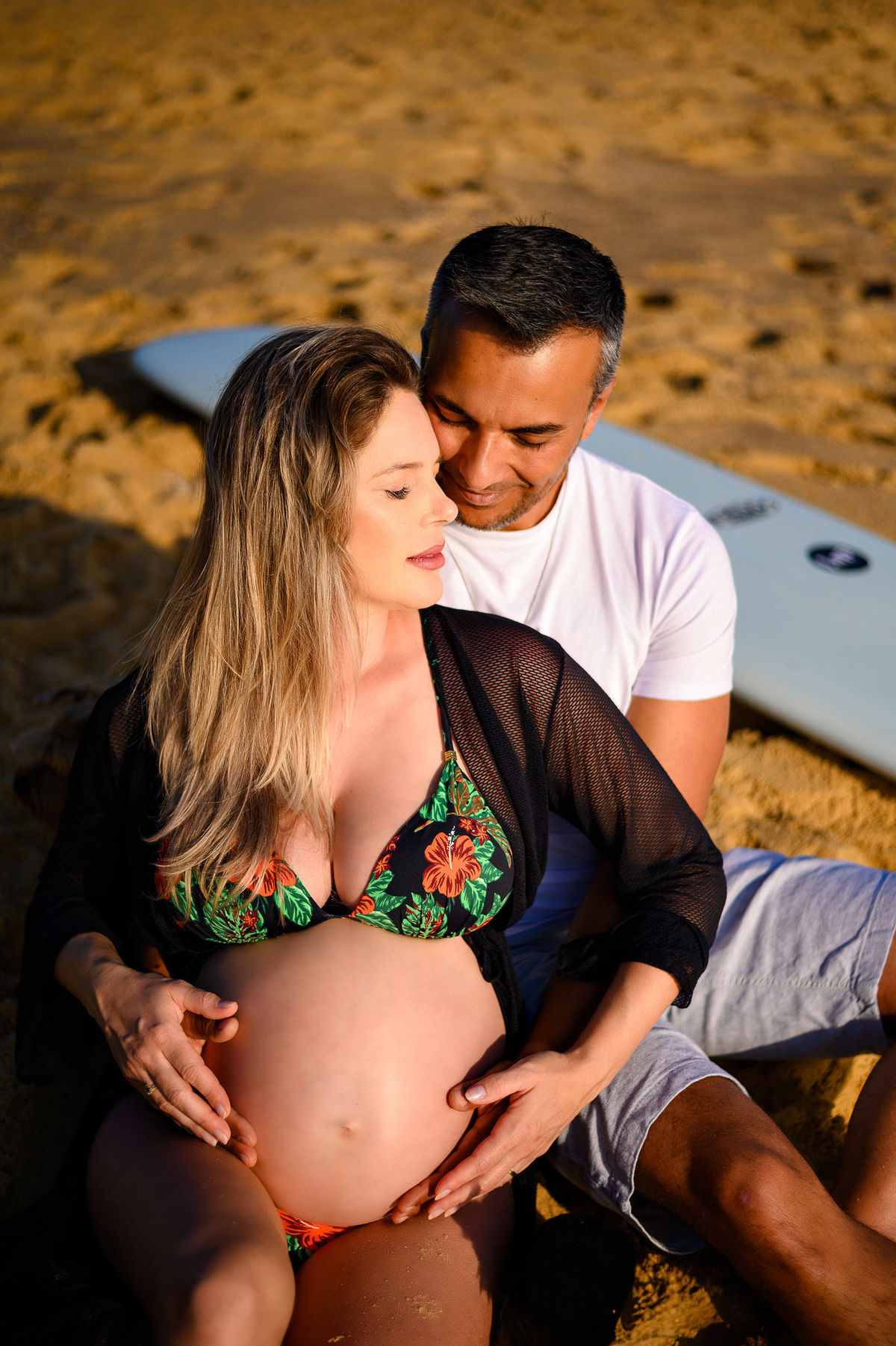 Casal em ensaio gestante na praia ao amanhecer em Balneário Camboriú com prancha de surf, em clima íntimo e natural.