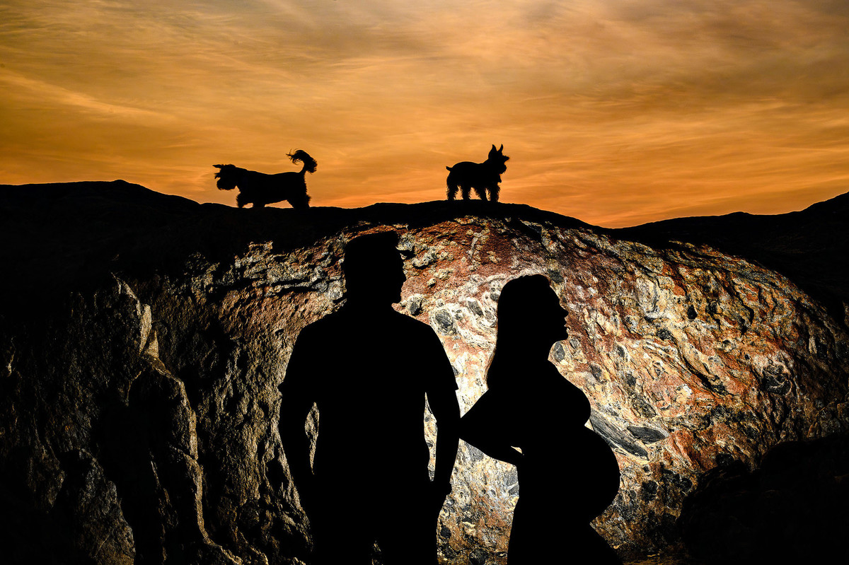 Gestante e companheiro em ensaio fotográfico na praia ao amanhecer em Balneário Camboriú com silhueta e pedras da praia