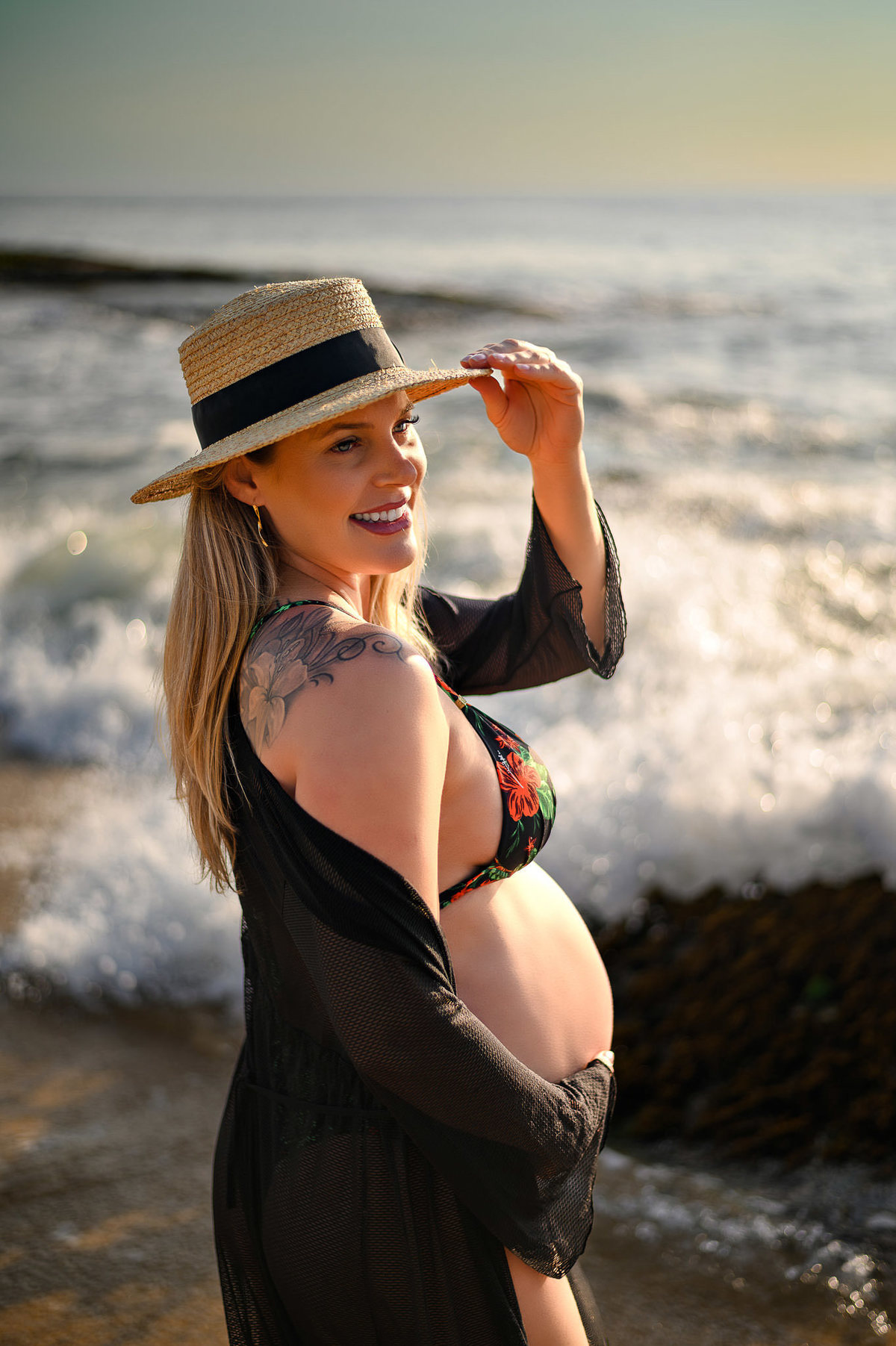 Gestante em ensaio fotográfico ao amanhecer na praia em Balneário Camboriú usando chapéu, com o mar ao fundo e luz suave do nascer do sol.