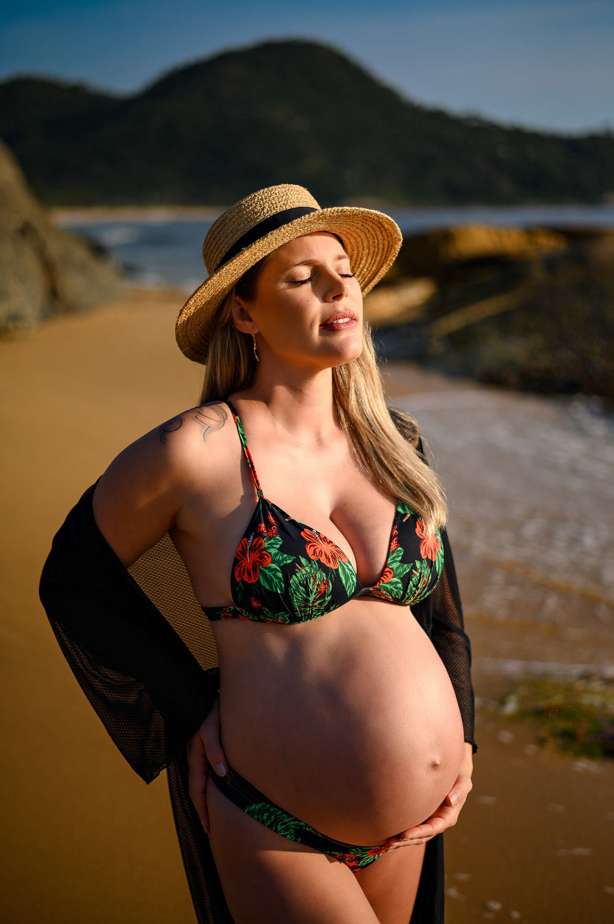 Gestante em ensaio fotográfico ao amanhecer na praia em Balneário Camboriú usando chapéu, com o mar ao fundo e luz suave do nascer do sol.
