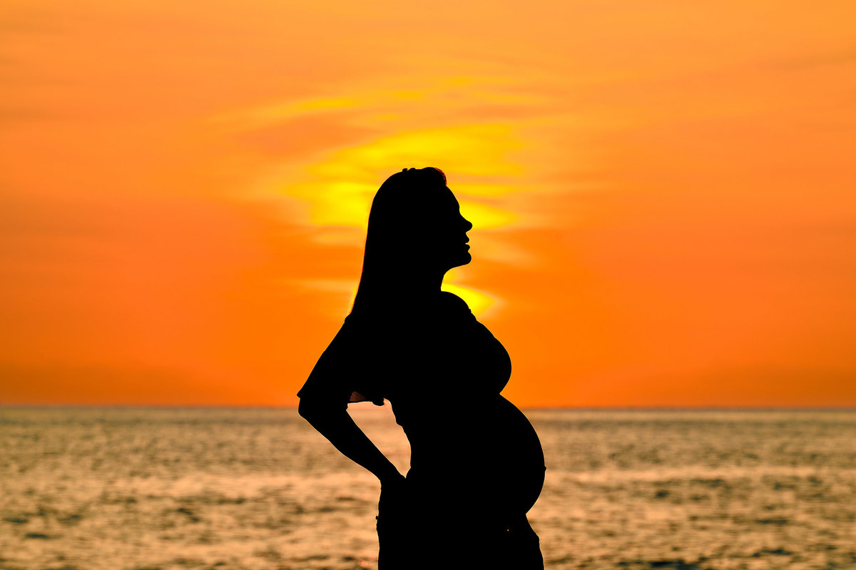 Silhueta de casal em ensaio gestante ao amanhecer na praia em Balneário Camboriú, com o sol nascendo ao fundo e o mar em contraluz.