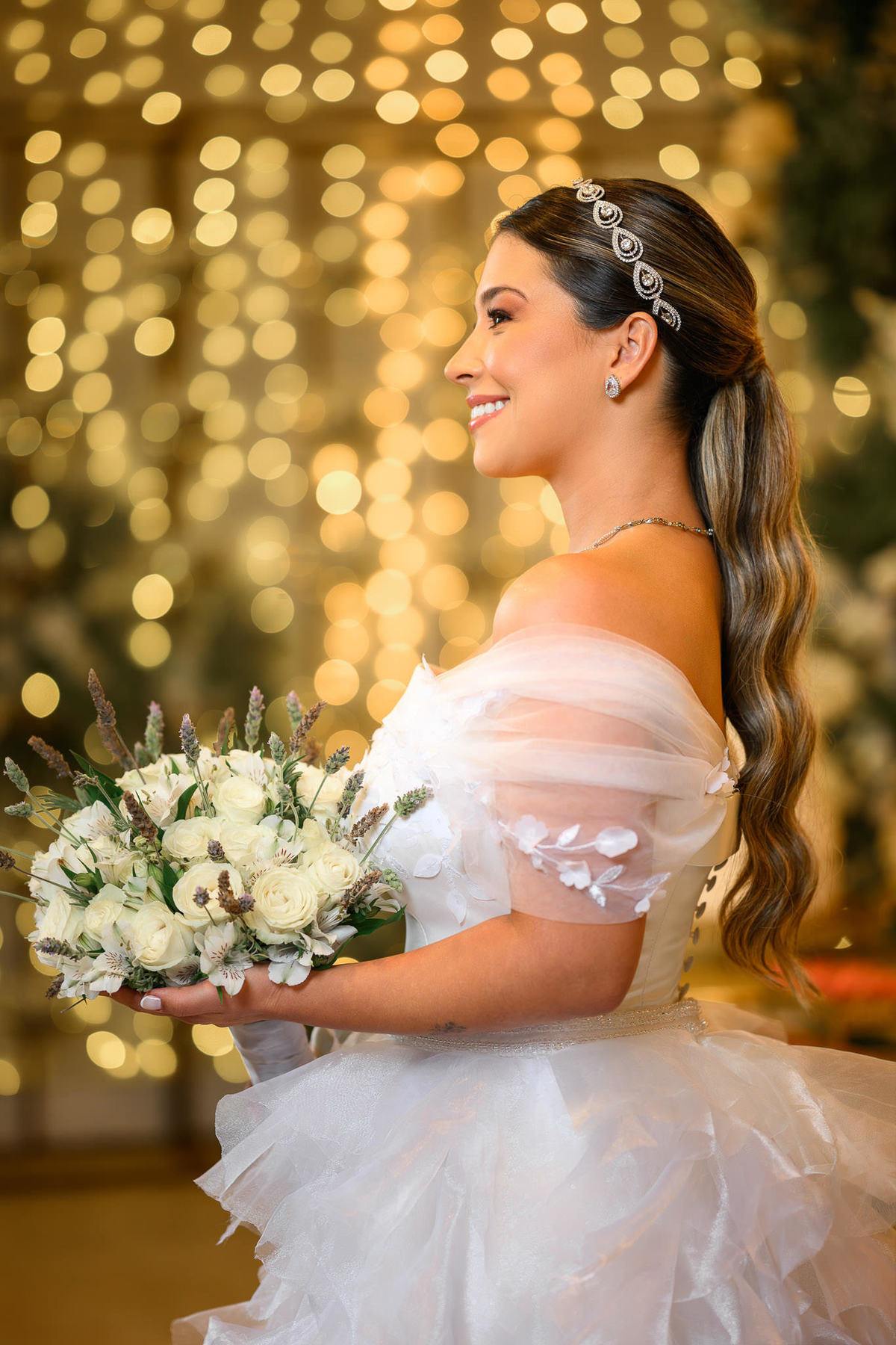 Noiva de perfil segura buquê branco diante de luzes douradas.