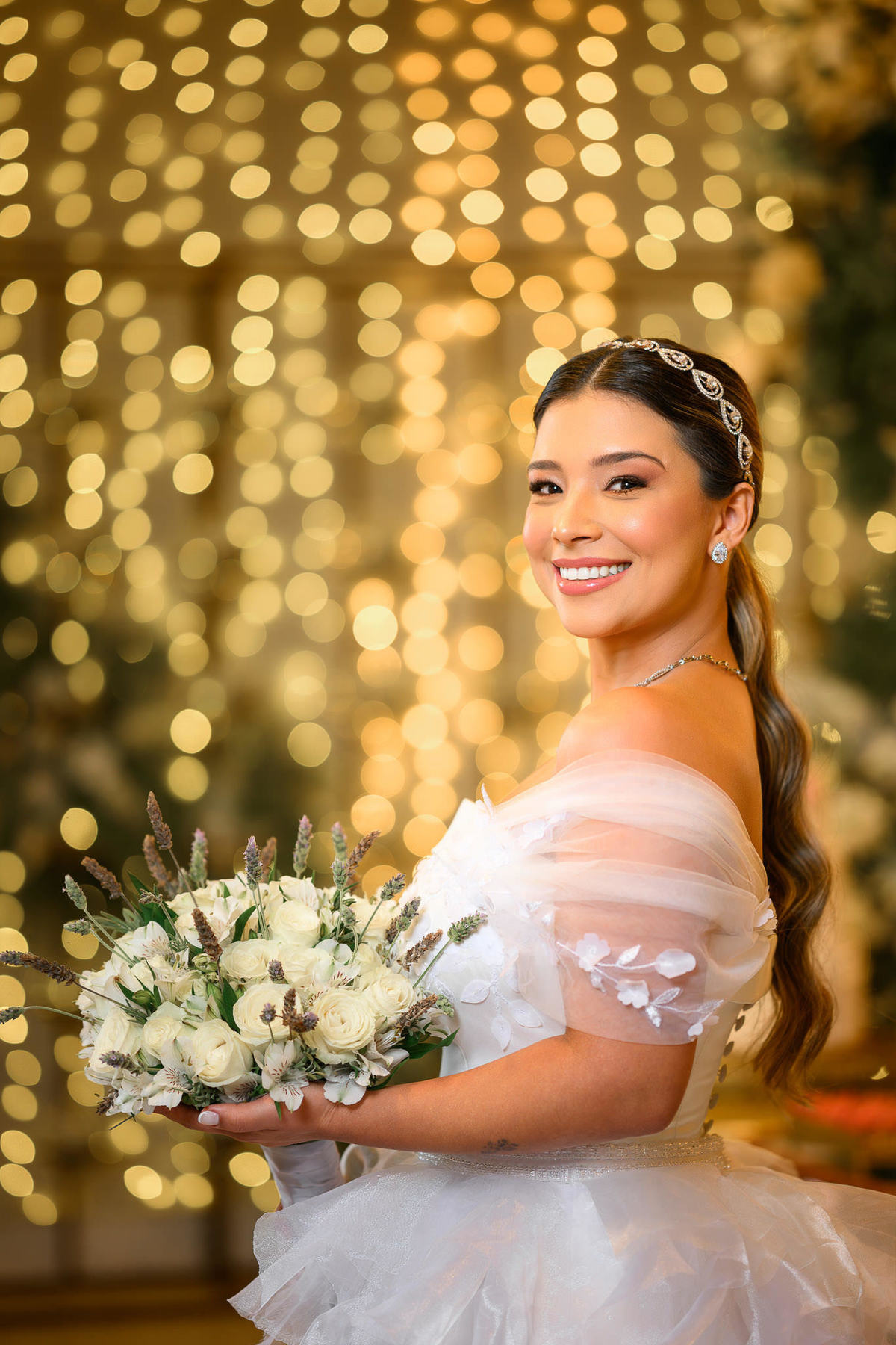 Noiva sorri para a câmera segurando o buquê diante da decoração iluminada.