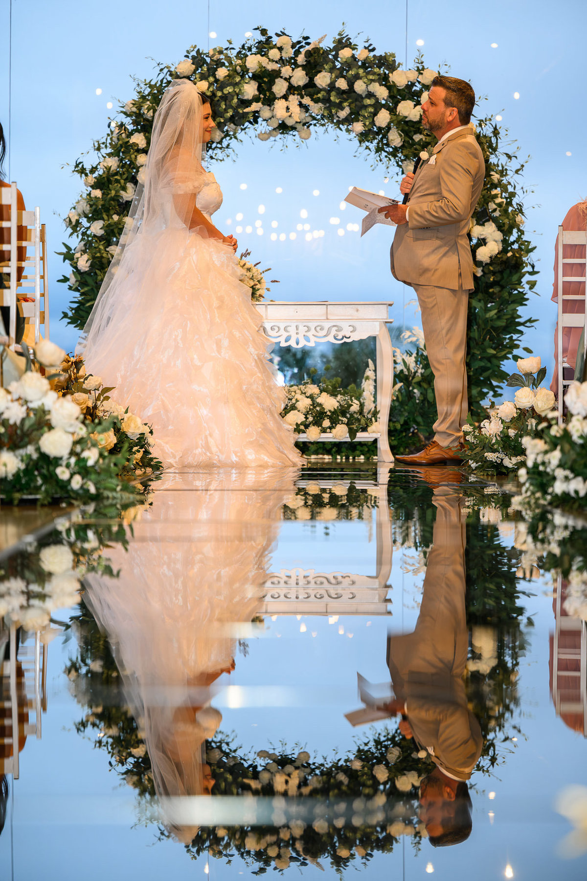 Noivos diante do arco floral refletidos no corredor espelhado da cerimônia no Indaiá Eventos Itapema