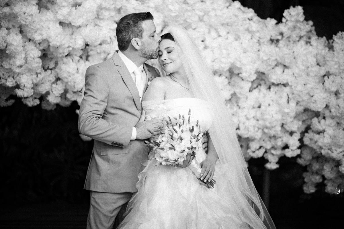 Noivo beija a testa da noiva entre flores brancas em fotografia em preto e branco.