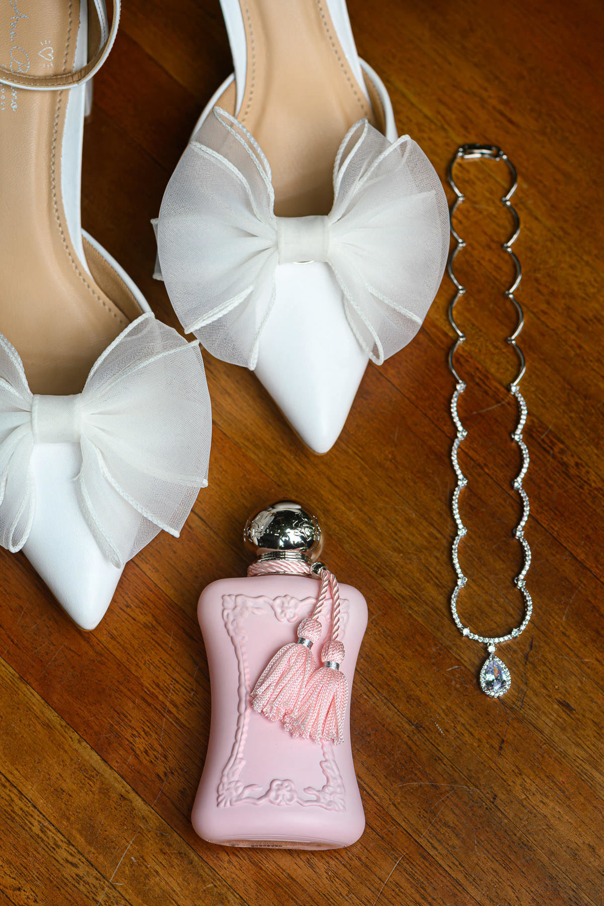 Sapatos brancos com laços, perfume rosa e colar da noiva sobre uma mesa de madeira.