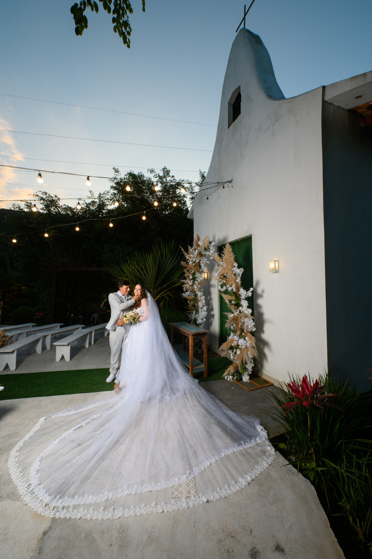Casal diante da capela com o longo véu aberto no chão