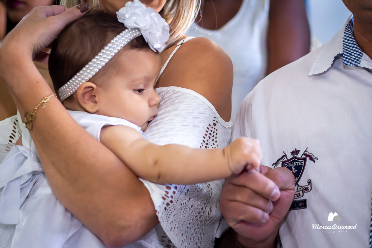 manina que será batizada com um vestido branco, uma tiara no cabelo, sendo carregada pela madrinha e segurando o dedo do padrinho e olhando pala o dedo que está segurando dentro da igreja que está sendo batizada