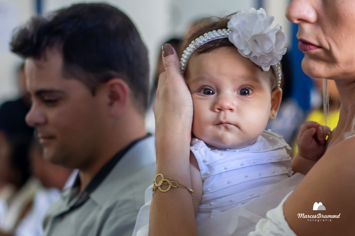 batizado da Lívia Manuela, com um vestido branco, olhando para câmera, sendo carregada pela madrinha e com o pai desfocado no fundo dentro da igreja a qual está sendo batizada