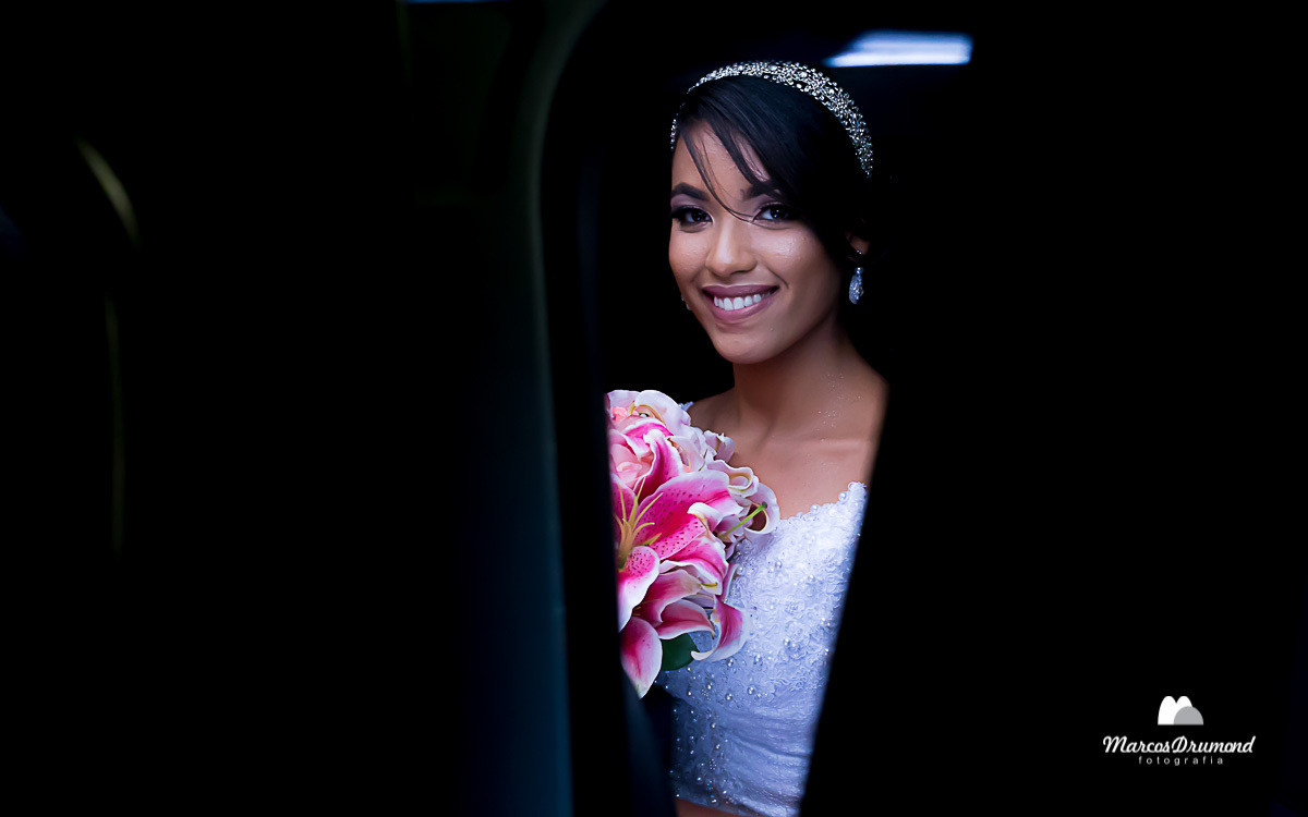 Noiva dentro do carro com seu vestido branco e o buquê nas mãos arco no cabelo e brincos nas orelas sorrindo e sendo fotografada pelo fotógrafo de casamentos Marcos na em São Gonçalo do Rio Abaixo/MG  Drumond