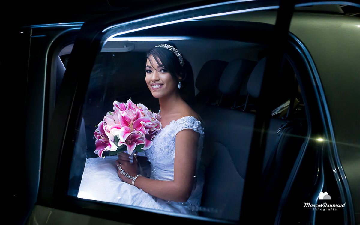Noiva dentro do carro com seu vestido branco e o buquê nas mãos arco no cabelo e brincos nas orelas sorrindo e sendo fotografada pelo fotógrafo de casamento Marcos Drumond minutos antes do início da cerimônia