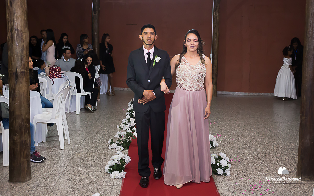 Noivo entrando para casar e sendo acompanhado por sua mãe, onde o mesmo está com o terno preto e sua mãe com um vestido longo na cor clara e sorrindo. O casamento foi em um salão de festa em São Gonçalo do Rio Abaixo no Estado de Minas Gerais