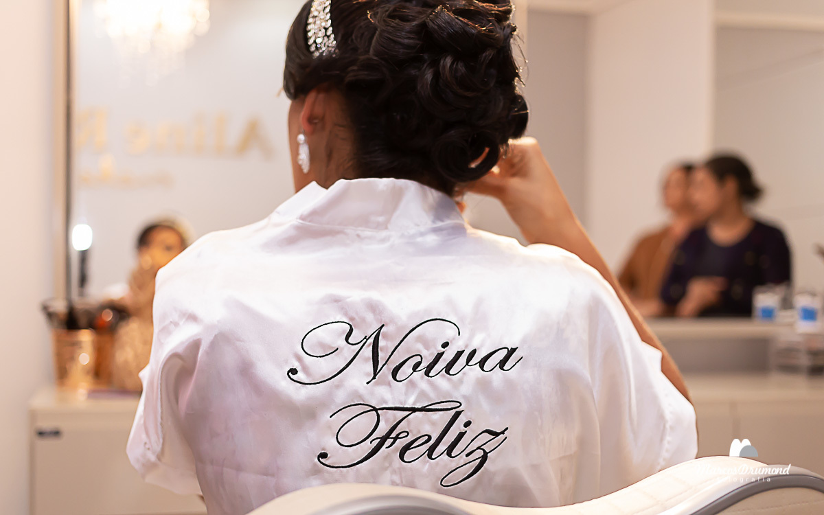 Noiva no salão de costas arrumando o brinco na frente do espelho com o cabelo preso e um arco no cabelo muito empolgada e sendo fotografada pelo fotógrafo Marcos Drumond