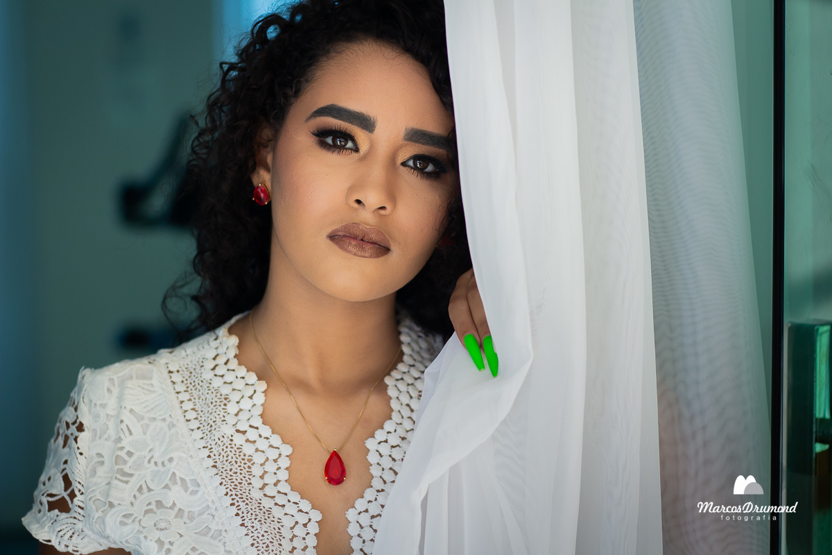 a modelo de pé vestida com uma blusa branca, brincos e colar com pedras vermelhas e unhas verdes na porta de vidro  segurando a cortina