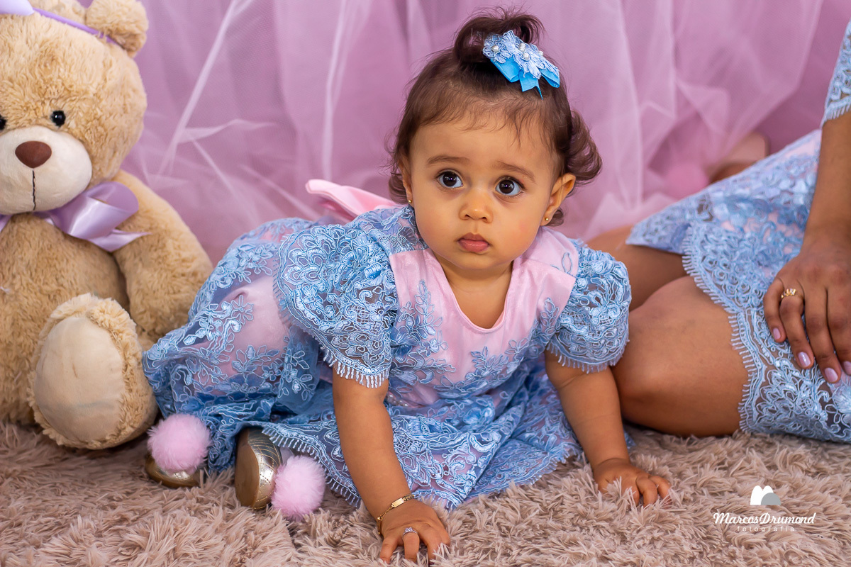 Foto colorida da aniversariante sentada ao lado da mesa de bolo olhando para frente