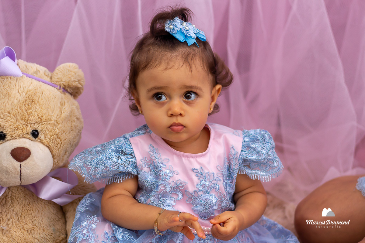Fotografia da aniversariante de 1 ano olhando para frente e mexendo no dedinho, com uma pulseira no bracinho e um anelzinho no dedo