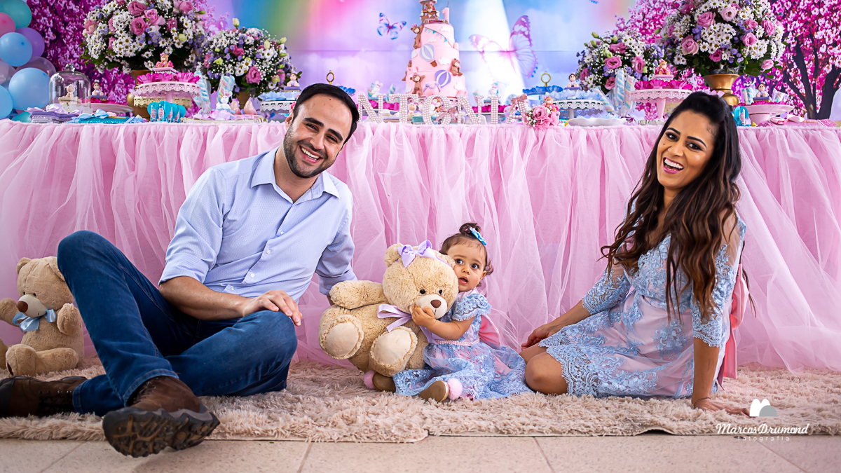 Fotografia de pai e da mãe da aniversariante de um ano segurando um ursinho e todos sentados perto da mesa de bolo olhando para frente e sorrindo
