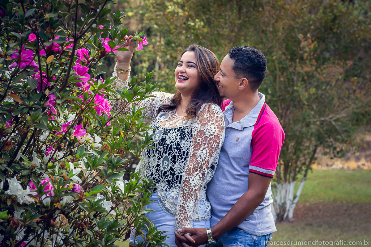 Pré casamento Carol e Varlei em Itabira/MG, onde a noiva esta sorrindo e pegando uma flor e o noivo está abraçado com ela segurando em sua mão e falando ao seu ouvido. O ensaio foi feito no sítio da cachoeira e Itabira/MG