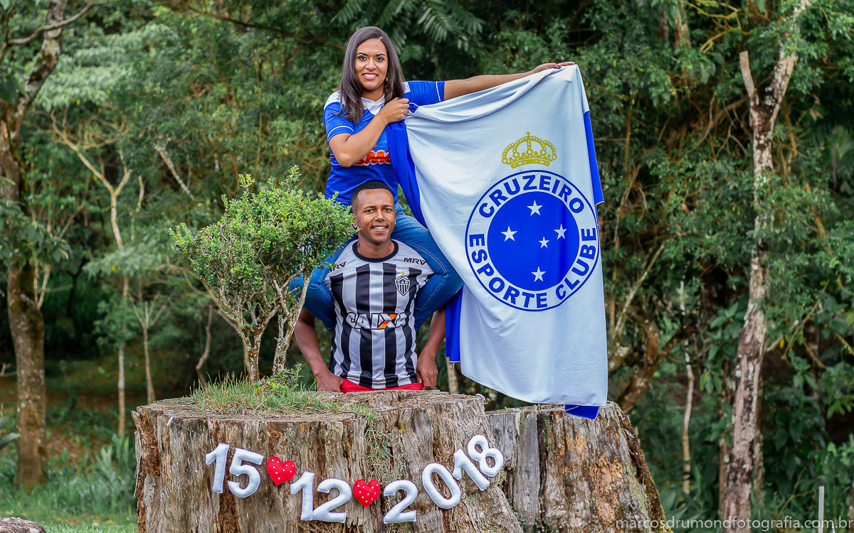 Fotografia de pré casamento onde a noiva está com a camisa do cruzeiro segurando uma camisa do cruzeiro sentada no ombro do noivo que está com uma camisa do galo e ambos estão olhando para a câmera