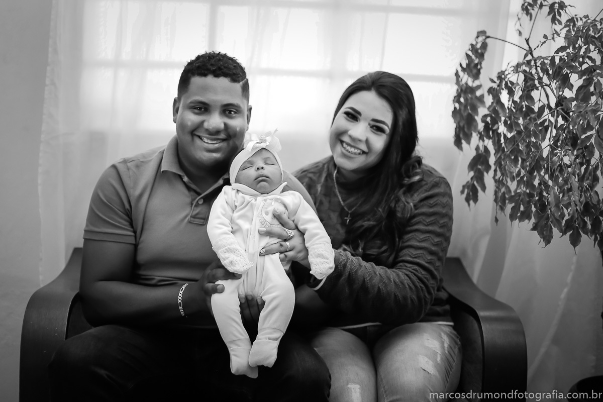 Fotografia em preto e branco feita na casa dos clientes, onde os pais estão segurando a menina de um ano que está usando uma roupa branca olhando para a câmera e sorrindo, eles estão sentados  no sofá na sala da casa, ao lado do sofá há algumas flores 