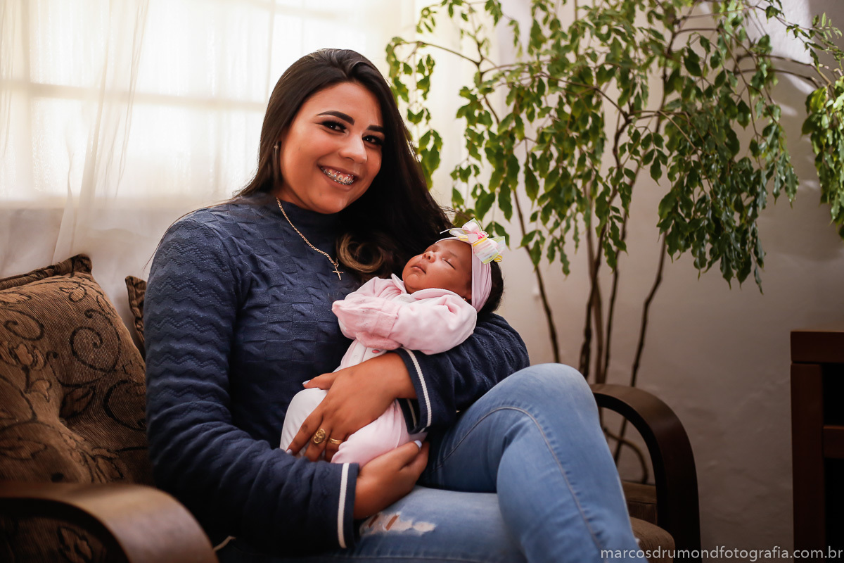 Fotografia mãe e filha sentada no sofá, onde a mãe está segurado a filha no colo olhando para a câmera e sorrindo