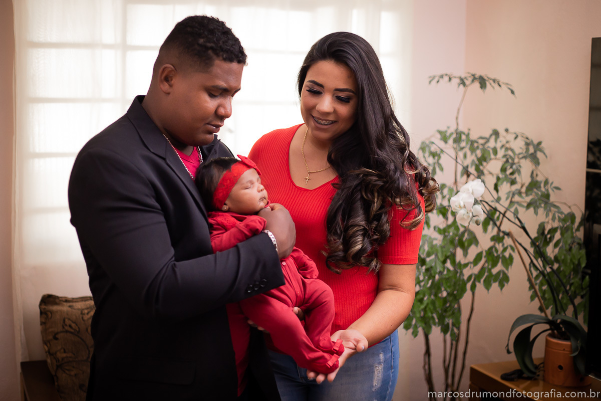 Sessão fotográfica de família realizada na casa dos clientes na cidade de Itabira/MG, onde o pai da menina está com um blazer preto, uma camisa vermelha, uma pulseira e um cordão prata, a mãe está de blusa vermelha e a criança vestida de vermelho também 
