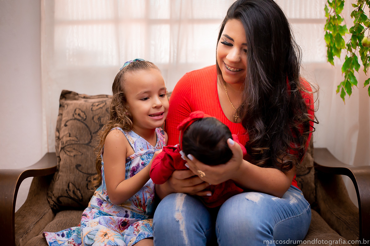Fotografia de família na casa dos clientes, a mãe está segurando a menina de um ano nos braços sentada no sofá, a mãe e a criança estão usando roupas vermelhas a mãe está com uma calça jeans clara e a priminha está sentada do lado