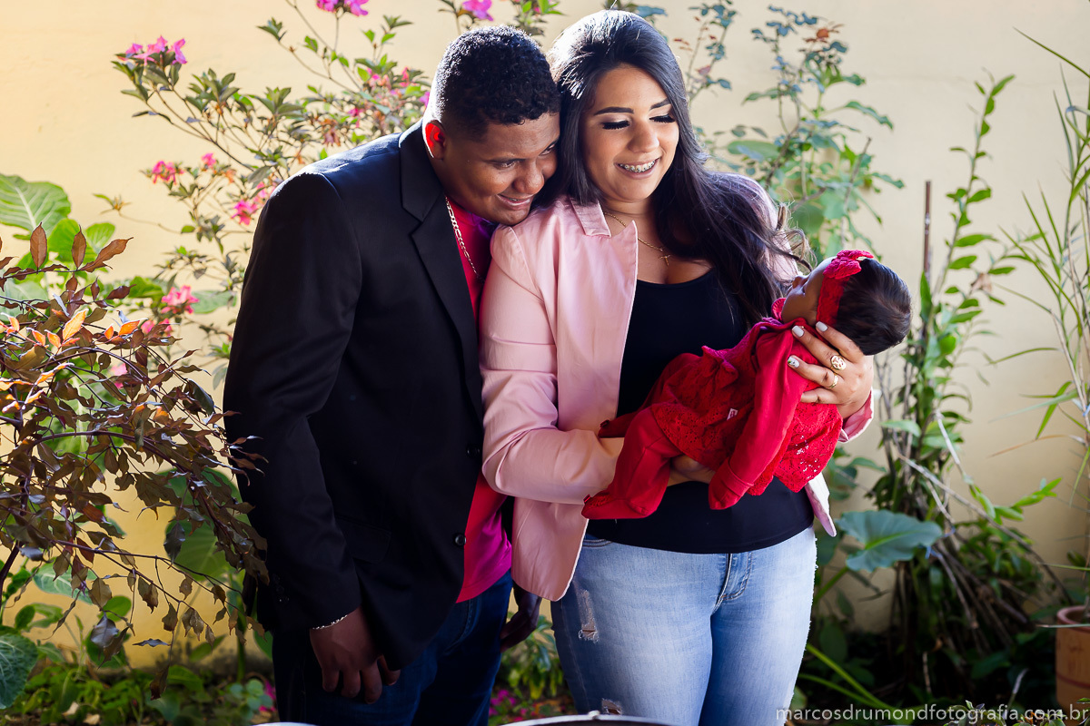 Fotografia dos pais segurando a menina de um ano de idade, eles estão olhando para ela, onde o pai está com o queixo apoiado no ombro da esposa e ambos estão sorrindo