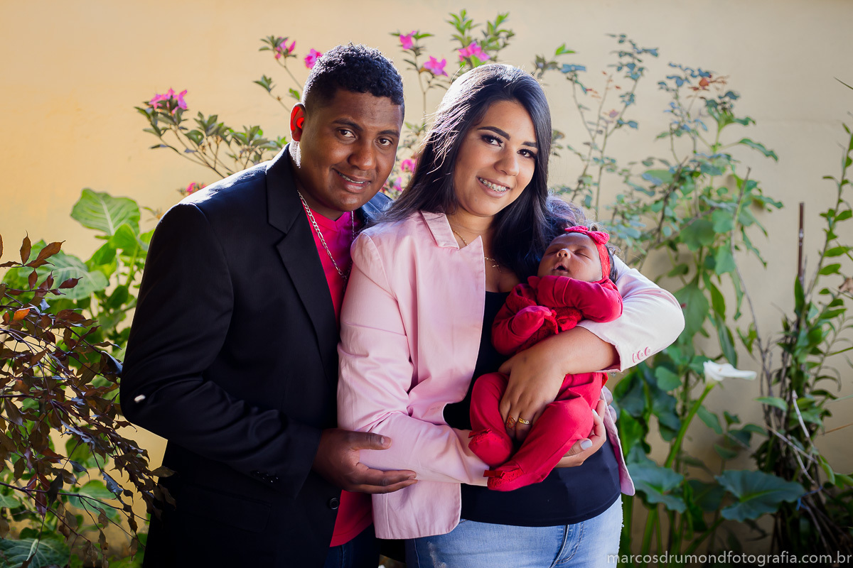 Fotografia feita na cidade de Itabira/MG n acasa da cliente, onde foram fotografados a mãe, o pai e a criança, a mãe está usando um blazer cor de rosa, blusa preta, calça jeans clara, o pai usa um blazer preto, camisa vermelha e calça jeans escura