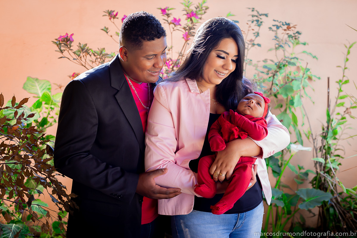 Fotografia feita na cidade de Itabira/MG n acasa da cliente, onde foram fotografados a mãe, o pai e a criança, a mãe e o pai estão olhando para a criança que está usando uma roupa vermelha, o pai está segurando no braço da mãe