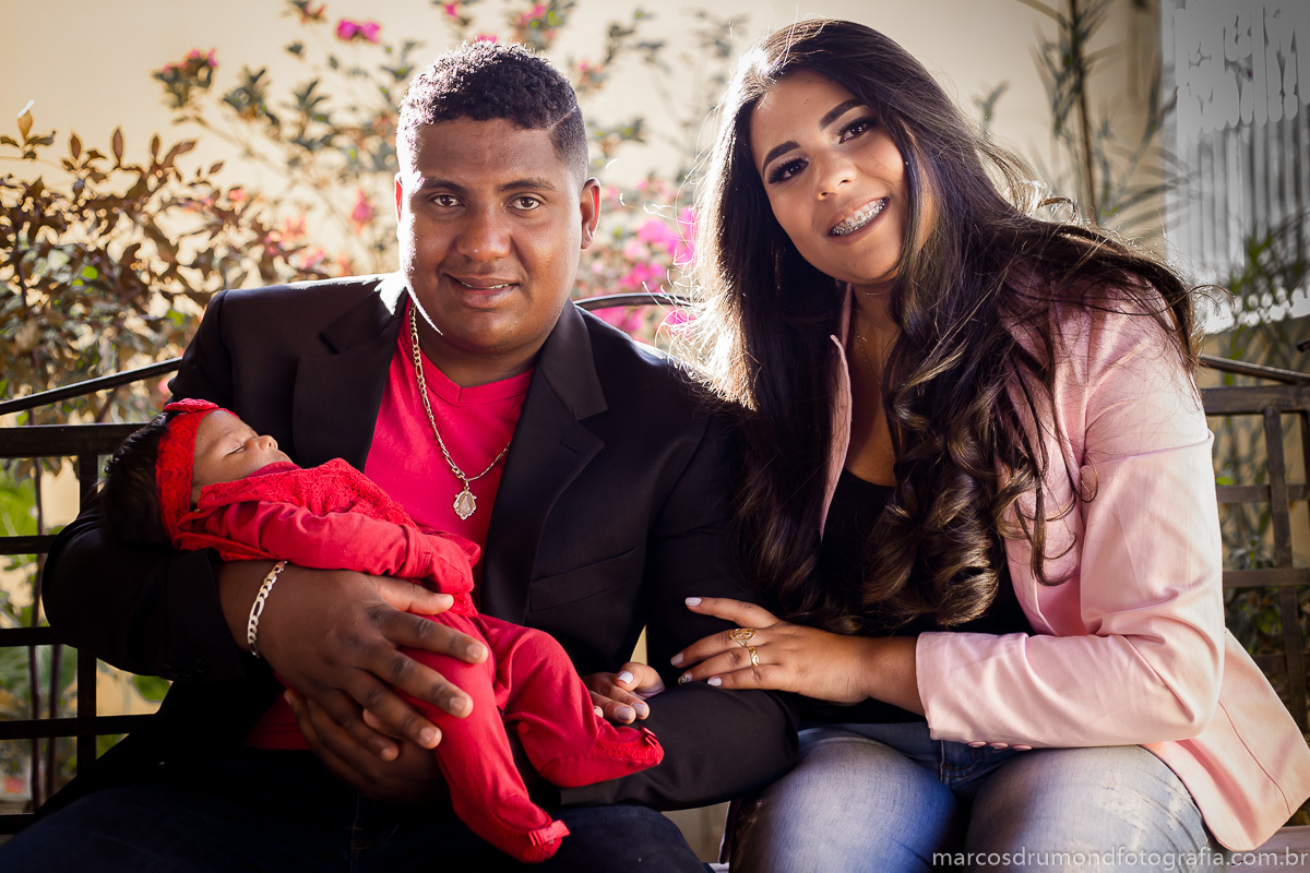 Ensaio fotográfico de família feito na cidade de Itabira/MG, onde a mãe, o pai estão olhando para a câmera e sorrindo, o pai está usando uma pulseira e um cordão prateados, um blazer preto, uma camisa vermelha, a mãe está usando um blazer cor de rosa