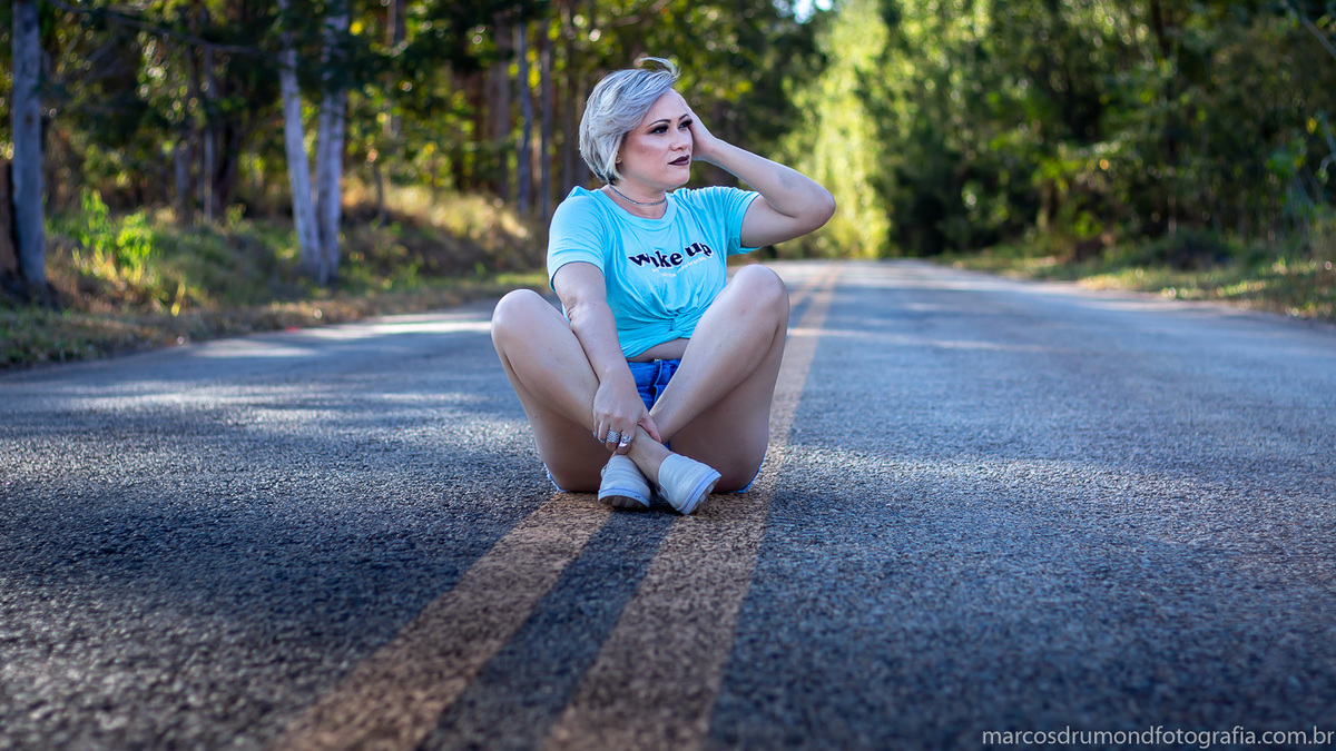 Ensaio fotográfico feminino externo onde a modelo está sentada no asfalto de bermuda jeans, tênis branco, camisa verde clara, cabelos loiros curtos com a mão na cabeça olhando para o lado