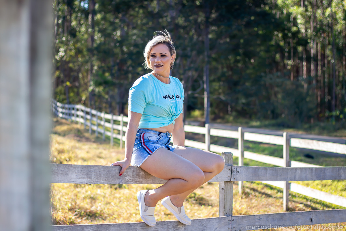 Ensaio fotográfico feminino durante o dia com a modelo sendo fotografada sentada em uma cerca e olhando pala o lado, a modelo está ousando um camisa verde clara, bermuda jeans, sapatênis e está segurando na cerca em um ambiente de vegetação, o sol show