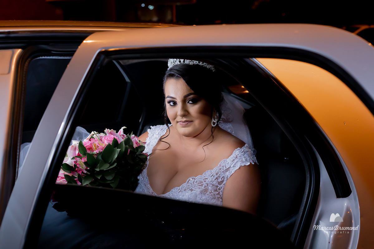 Fotografia da noiva dentro do carro chegando a igreja para o casamento, ela está com um belo vestido longo, branco, o buquê de flores nas mãos, brincos e olhando para fora, onde está sendo fotografada 