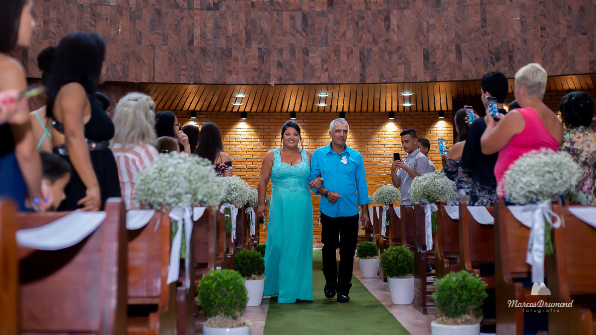 Fotografia colorida onde a mão da noiva está entrando na igreja com o pai do noivo, ela está usando um vestido longo na cor azul e sorrindo e pai do noivo está usando uma camisa azul manga longa e uma calça preta