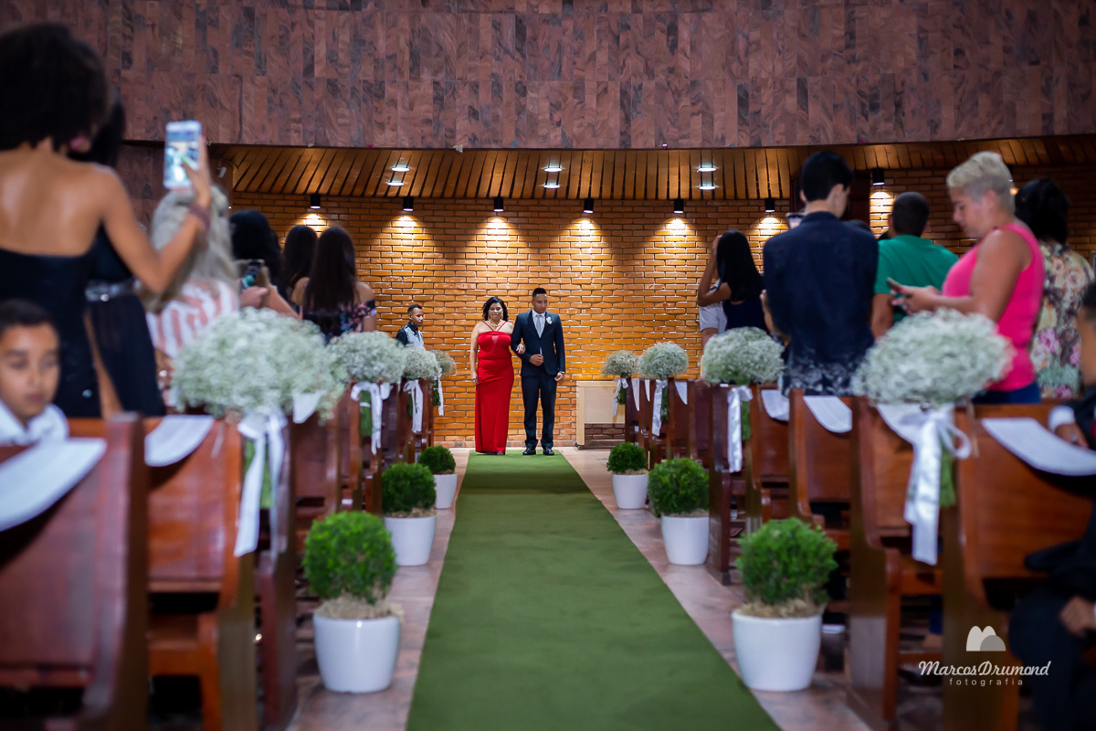 Fotografia colorida do casamento de Carol e Varlei, onde a mãe do noivo está vestida com um vestido longo na cor vermelha e entrando na igreja com o noivo seu filho que está com um terno na cor preta