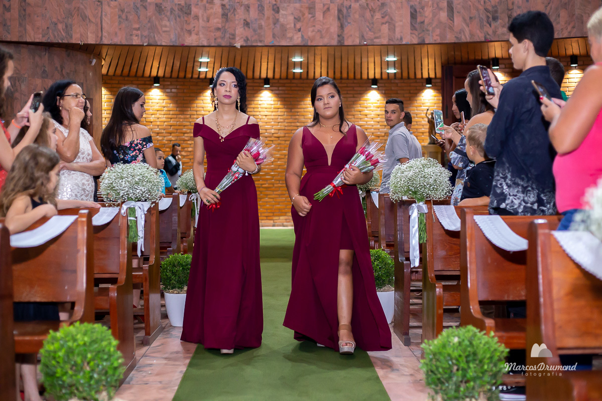 Damas entrando a igreja para o casamento de Carol e Varlei, elas estão usando vestidos na cor vermelha e segurando flores, olhando para frente e sendo registradas pelos fotógrafos que estão fotografando o casamento 