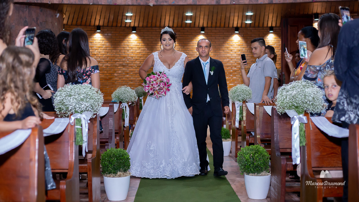 Fotografia colorida onde a noiva está entrando a igreja com o pai, ela está usando um vestido na cor branco e está segurando um buquê de flores, o pai está usando um terno na cor preto e ambos estão olhando para frente e sorrindo