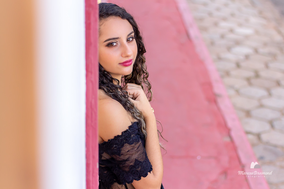 Fotografia colorida feminina urbana, onde a modelo está com as mãos próximo ao pescoço e olhando para a câmera, a foto foi feita em uma composição na parede de uma casa